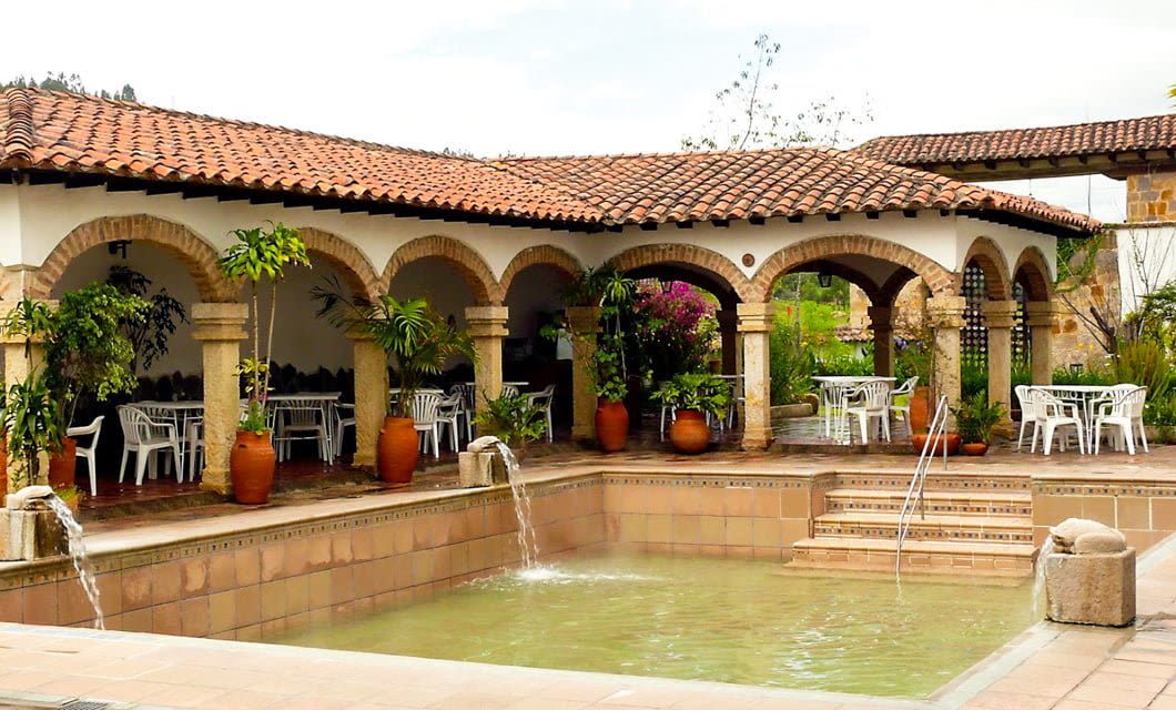 A courtyard in Boyaca, Colombia, with a terracotta-tiled roof, arched columns, potted plants, and white patio tables surrounds a small pool with water features—an inviting stop on your tour.