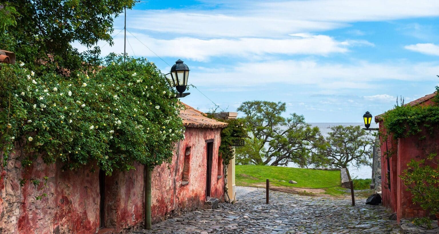 A narrow cobblestone street lined with old, weathered buildings and flowering plants leads to a grassy area overlooking a river under a bright, partly cloudy sky—an inviting scene often featured on Uruguay tours.