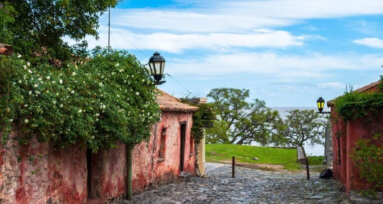 A narrow cobblestone street lined with old, weathered buildings and flowering plants leads to a grassy area overlooking a river under a bright, partly cloudy sky—an inviting scene often featured on Uruguay tours.