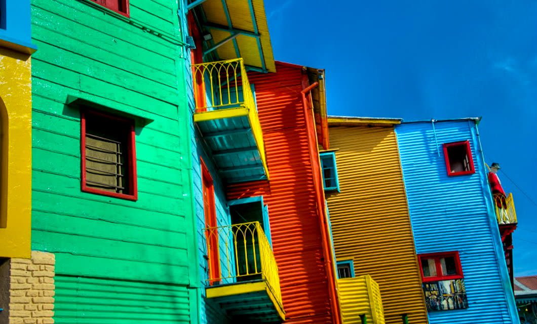 Brightly painted buildings in green, yellow, orange, and blue with balconies, under a clear blue sky. The vibrant colors evoke the lively spirit found along a Railway Journey like the Tucuman-Buenos Aires Railway.
