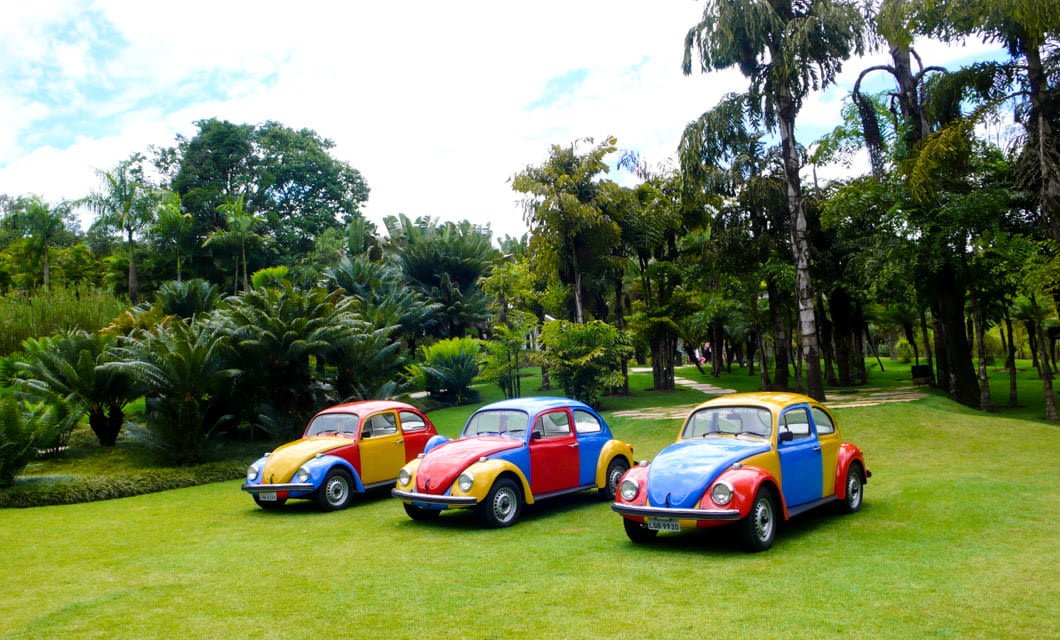 Three vintage Volkswagen Beetles, each painted in bright primary colors, are parked side by side on a lush green lawn surrounded by tropical trees—evoking the vibrant spirit of a Brazil Tour through its colonial gems and historic cities.