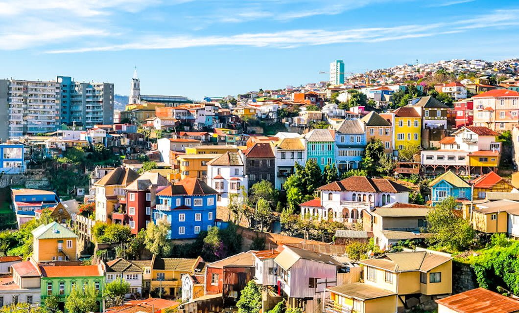 Colorful houses line the hillsides of a vibrant South American coastal city under a bright blue sky, with greenery, modern buildings, and the promise of an unforgettable wine tour in the background.