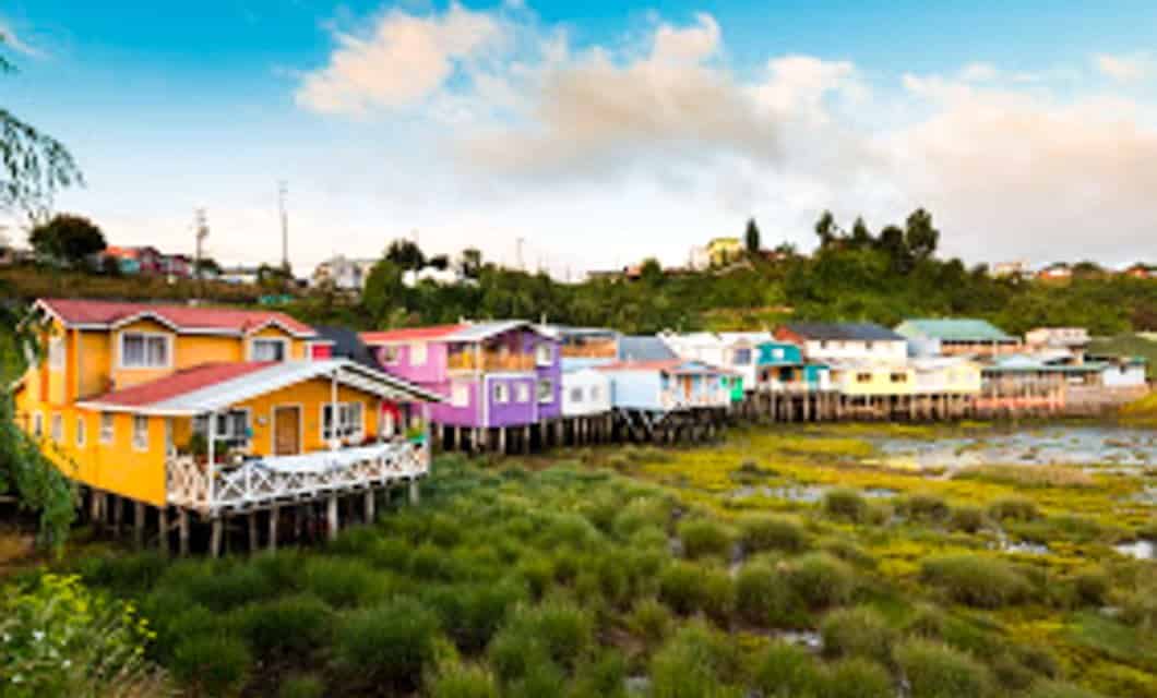 Colorful wooden houses on stilts line a grassy, marshy Chilean shoreline, with green hills in the background and a partly cloudy sky overhead.