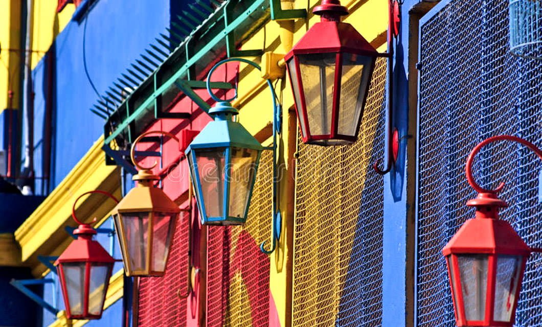 Colorful lanterns hang from a building painted in vivid blue, yellow, and red tones, with metal mesh over the windows—a bright, lively scene that captures the artistic urban vibe of a Buenos Aires tango tour.