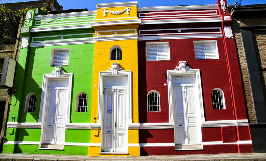 Three adjacent houses with identical architecture are painted in bright green, yellow, and red vertical stripes, each with a white door and white-framed windows, evoking the vibrant charm of scenes along the historic Tucuman-Buenos Aires Railway.