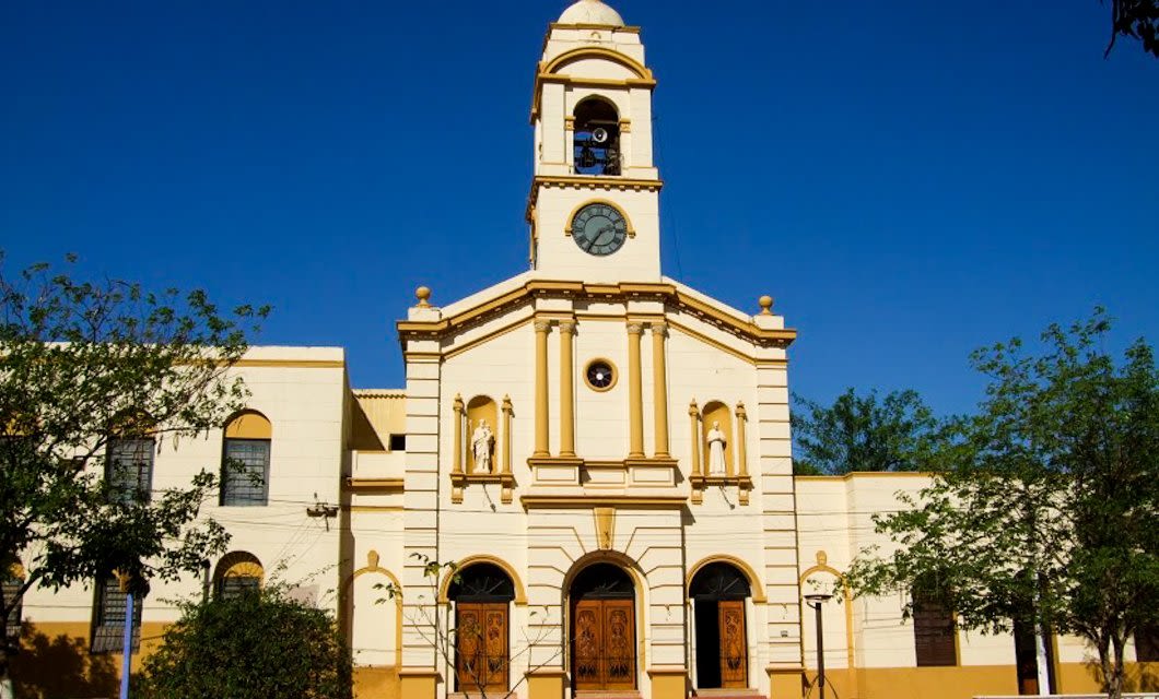 Yellow and white church with a central clock tower, arched wooden doors, and two statues in niches. Surrounded by trees, this charming spot in Paraguay is perfect to visit on your tour to Iguazu Falls, with a clear blue sky in the background.