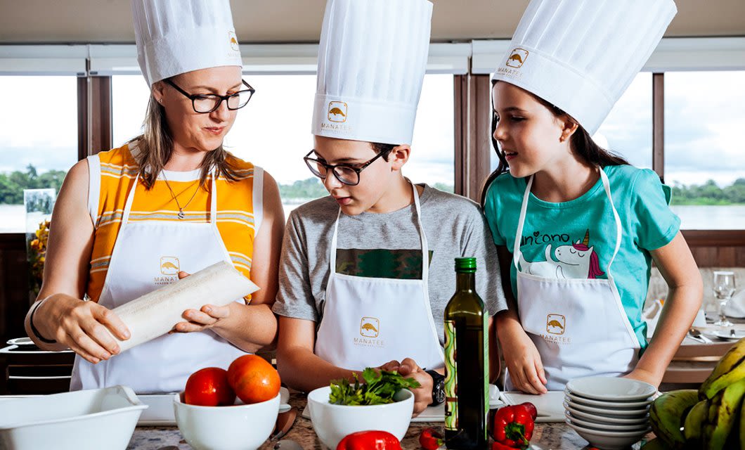 Three people wearing chef hats and aprons prepare food together in a bright kitchen. The woman holds a rolled wrap while the two children watch, dreaming of an Amazon Explorer adventure through the Ecuadorian Rainforest. Fresh veggies and olive oil are on the counter.