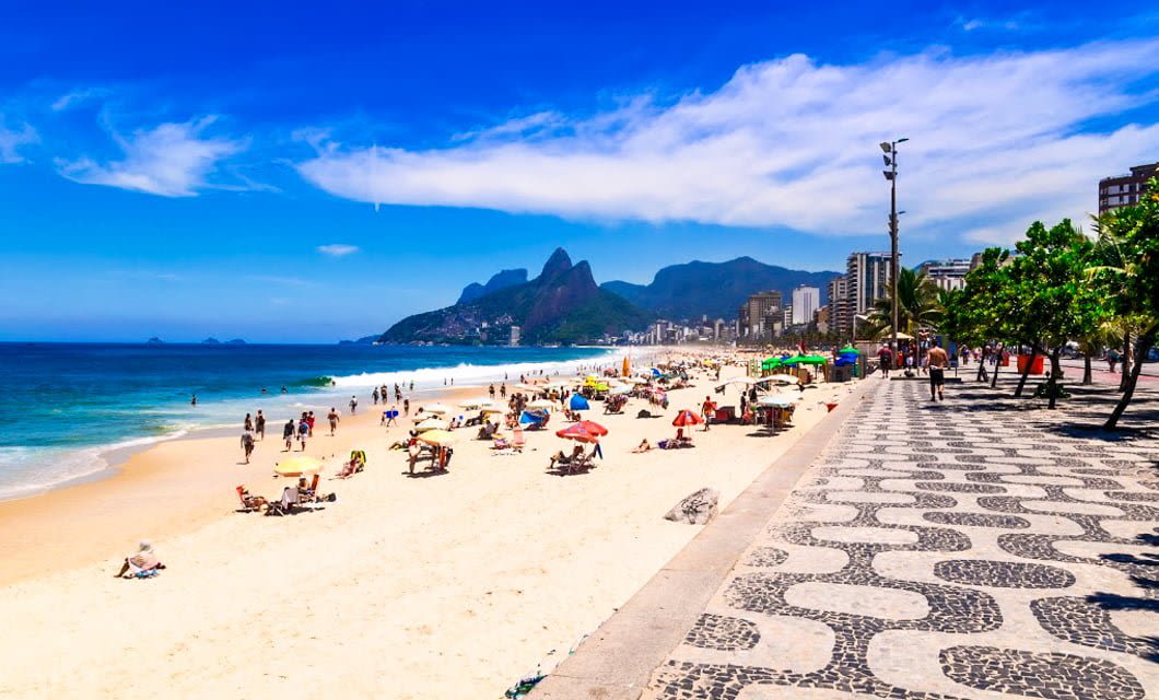 A sunny beach in Brazil with people sunbathing, a wavy-patterned promenade, palm trees, city buildings, and green mountains under a blue sky—perfect for your New Year's tour.