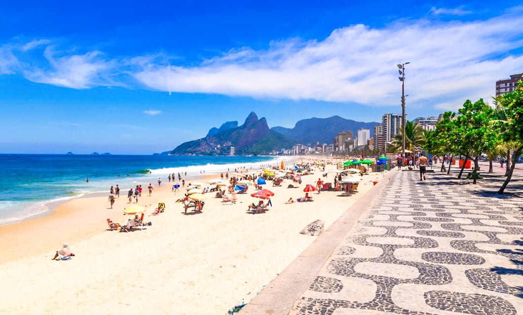 A sunny beach scene with people relaxing under umbrellas on sandy shores, clear blue ocean waves, a patterned sidewalk, palm trees, and city buildings with mountains in the background—perfect for a Brazil Tour.