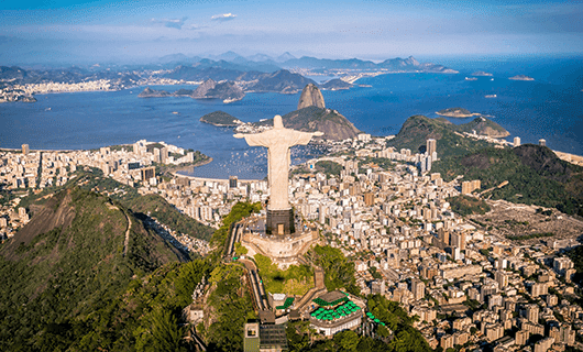 Aerial view of the Christ the Redeemer statue overlooking Rio de Janeiro, with urban buildings, mountains, beaches, and Guanabara Bay in the background.