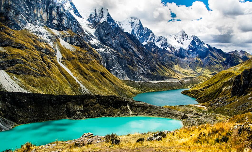 Turquoise lakes surrounded by steep, grassy slopes and dramatic snow-capped peaks define this scenic, remote Cordillera Blanca landscape near Huaraz, perfect for a breathtaking Peru trek under a bright, partly cloudy sky.