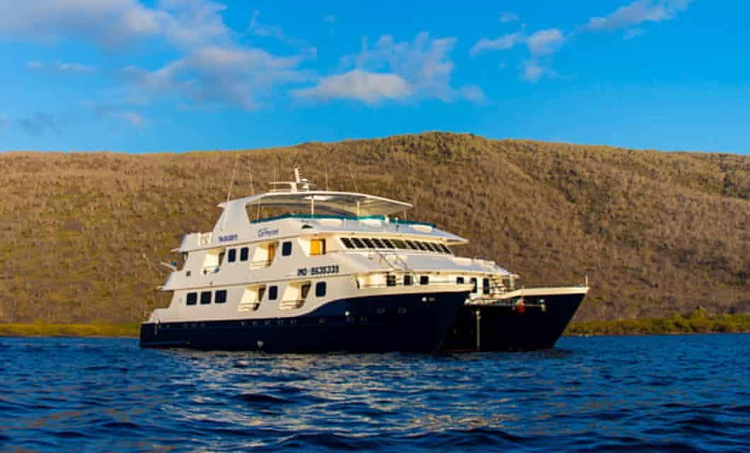 The M/C Cormorant, a large white and navy blue yacht, floats on calm blue water with a rocky, brown hillside and partly cloudy sky in the background—perfect for an unforgettable Galapagos cruise.