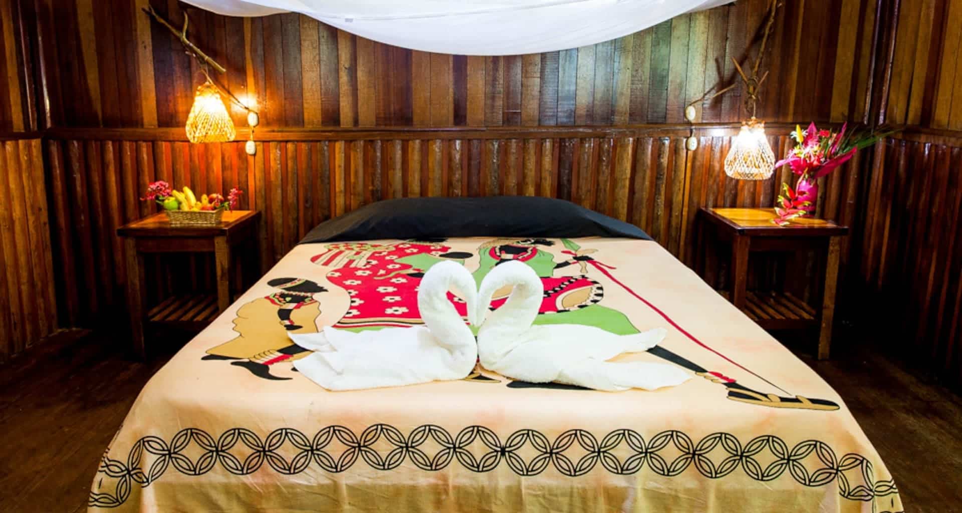 A cozy wooden cabin bedroom at Corto Maltes Lodge with a double bed, colorful patterned bedspread, towel swans on the bed, two bedside tables with flowers, and warm hanging lamps on each side.