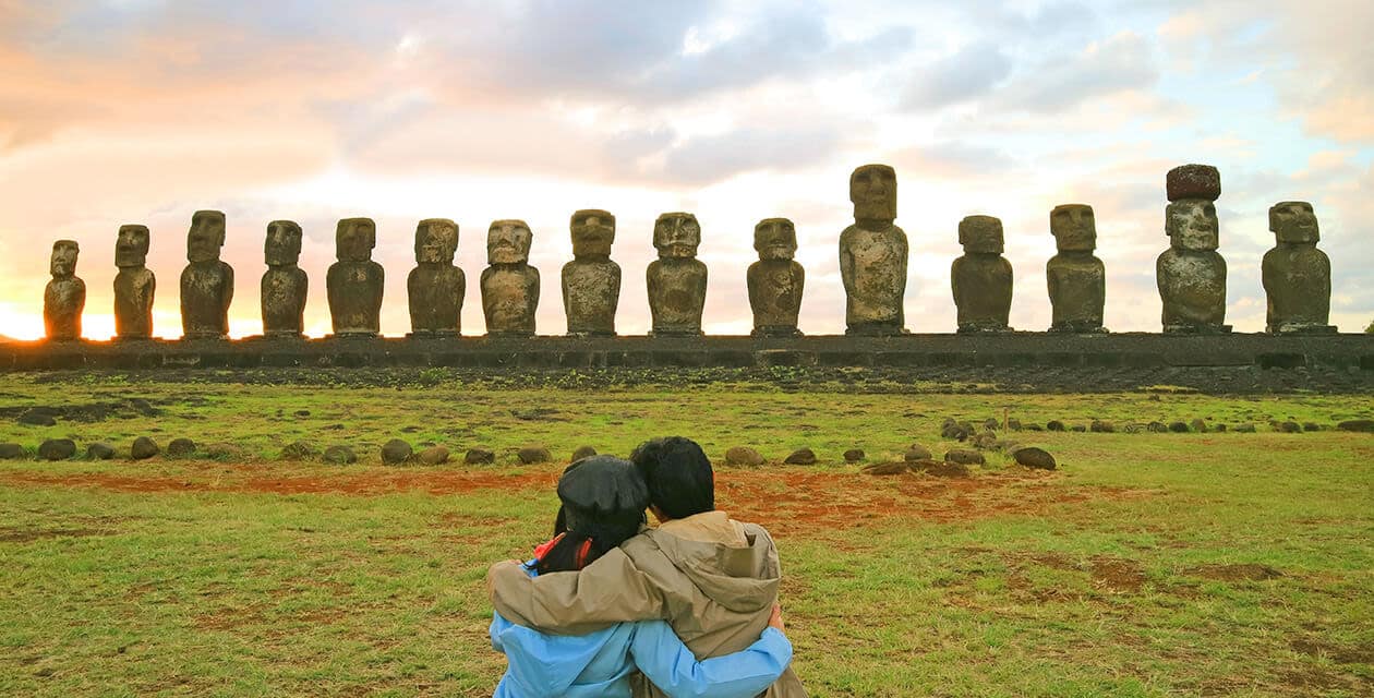 Two people sit on grass, arms around each other, facing a row of large stone moai statues on Easter Island at sunset. The sky is partly cloudy with warm sunlight illuminating the scene.
