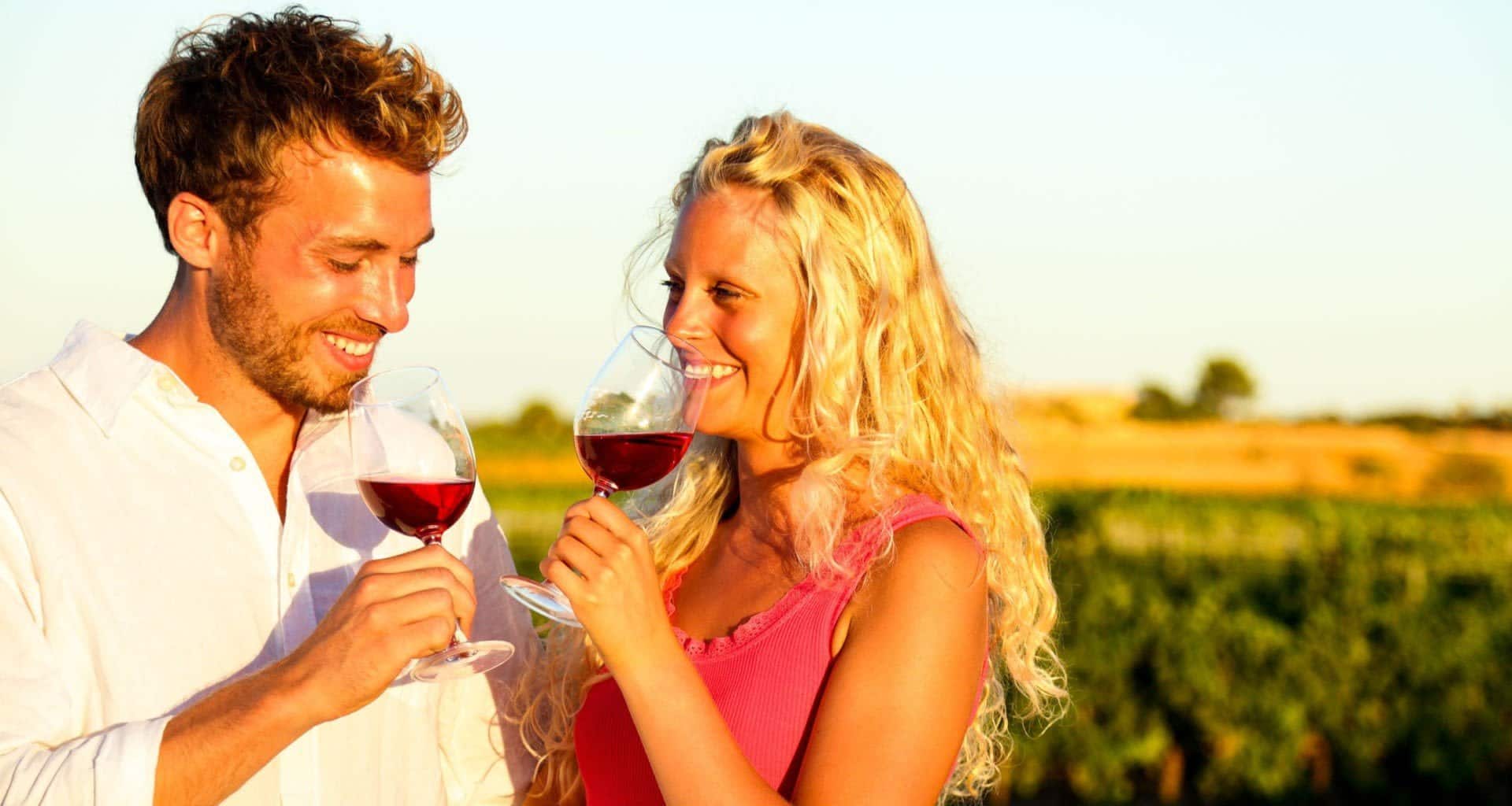 A smiling man and woman stand outdoors in a South American vineyard at sunset, holding glasses of red wine and looking at each other. The woman wears a pink dress, and the man wears a white shirt—perfect for a romantic wine tour.