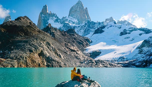 Two people sit on a rock by a turquoise lake, facing snow-capped mountains under a clear blue sky—an unforgettable family journey through rugged terrain and dramatic peaks.
