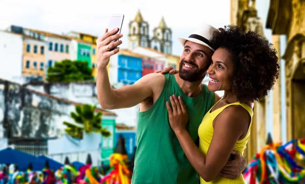 A smiling couple takes a selfie outdoors on their South America Tour, surrounded by colorful buildings and festive decorations. The man wears a hat and green tank top; the woman wears a yellow dress. Two church towers rise in the background.