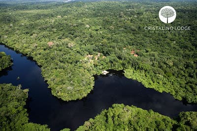 Aerial view of a lush green rainforest with a winding river and a small dock, capturing the true Amazonian experience. The Cristalino Lodge logo appears in the top right corner.