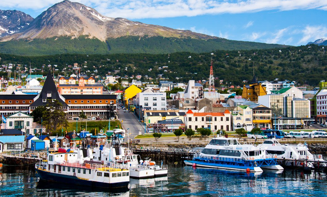 Colorful waterfront town with boats docked at a marina in Patagonia, backed by green hills and a tall, rugged mountain under a partly cloudy sky.