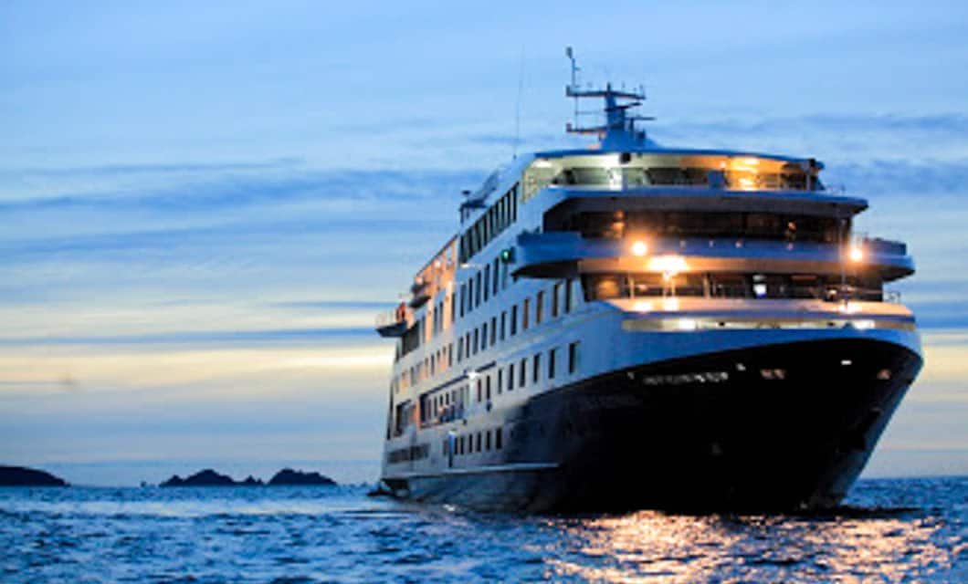 A large cruise ship sails on calm ocean waters at dusk, its windows aglow with light, as it journeys from Buenos Aires toward the scenic horizon of Tierra del Fuego.