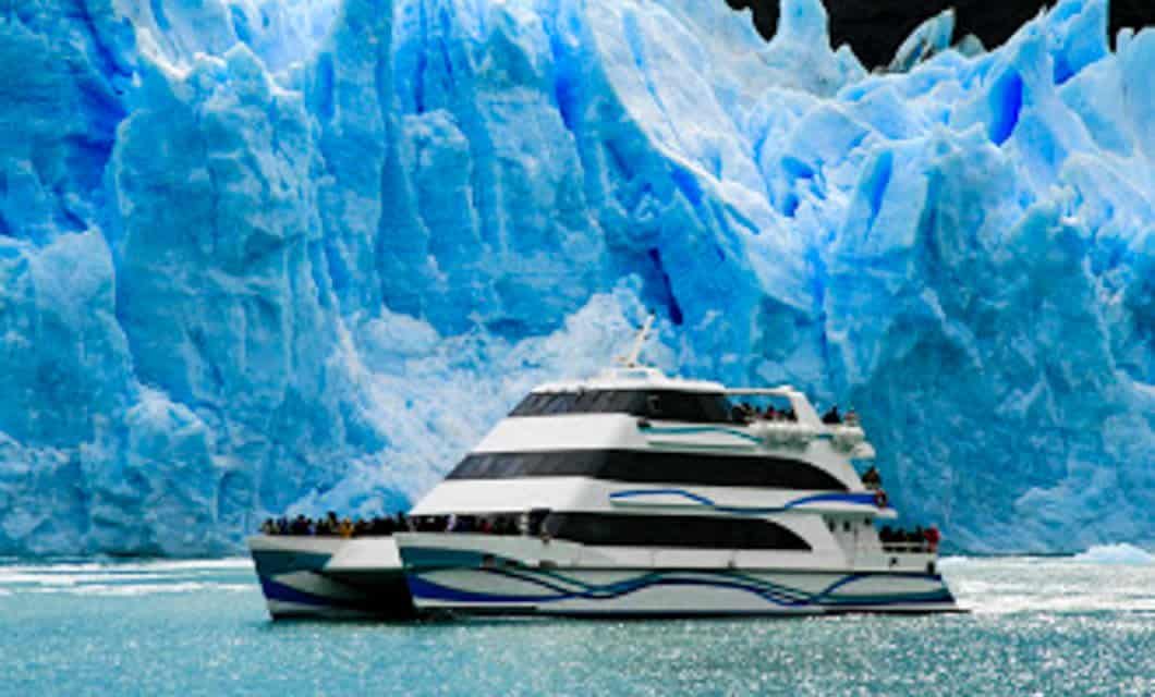 A large white and black tour boat sails close to a massive blue glacier in Argentina Patagonia, with numerous people on the decks observing the ice formation. The water is a vivid turquoise color.