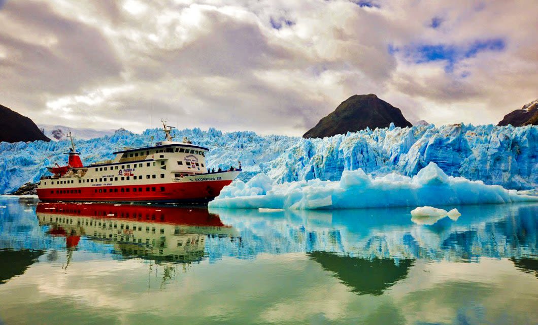 A red and white Skorpios Cruises ship sails on calm, reflective water near a massive blue glacier with snow-capped peaks in the Chilean Fjords of Patagonia, beneath a cloudy sky.
