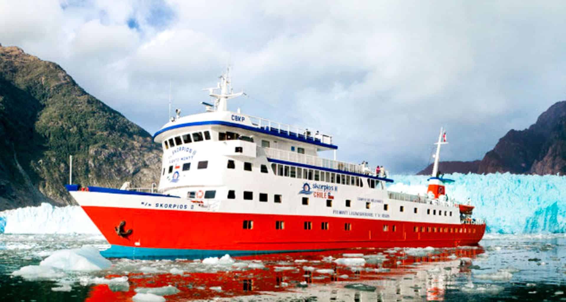 A bright red and white Skorpios Cruises ship sails through icy waters near a glacier in the Chilean Fjords, surrounded by floating ice and flanked by rugged, mountainous terrain under a partly cloudy sky.