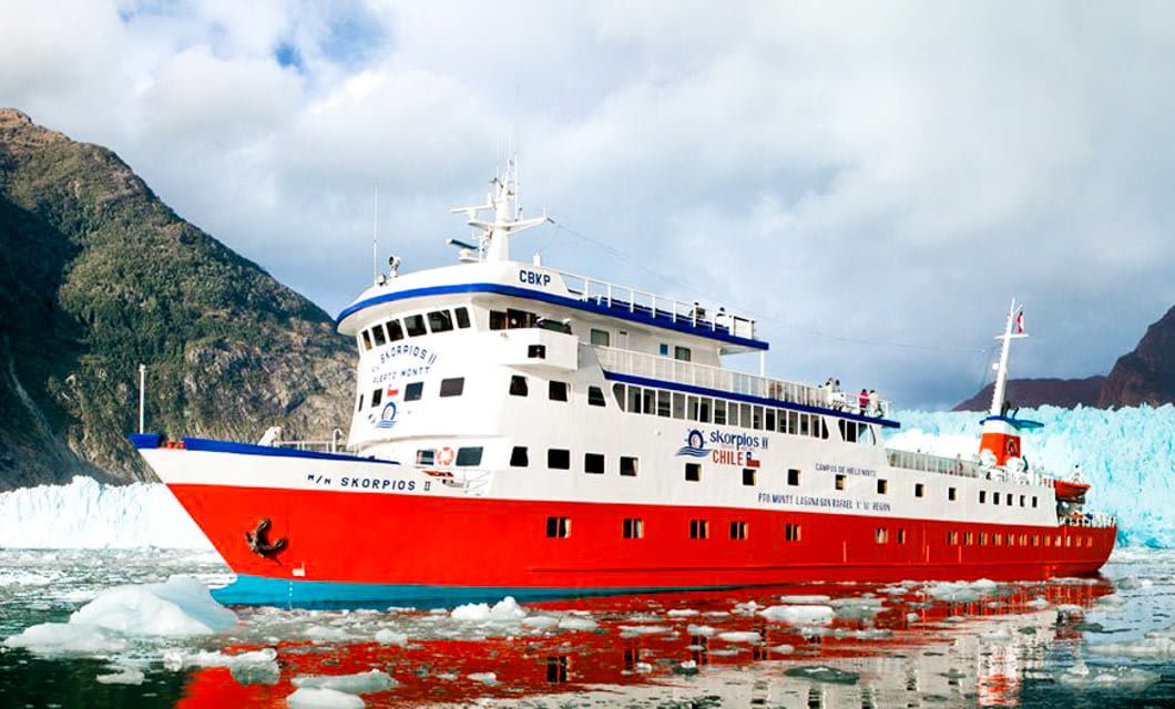 A large red and white Skorpios Cruises ship sails through the icy waters of the Chilean Fjords, with floating ice chunks, surrounded by majestic mountains and a glacier in Patagonia under a partly cloudy sky.