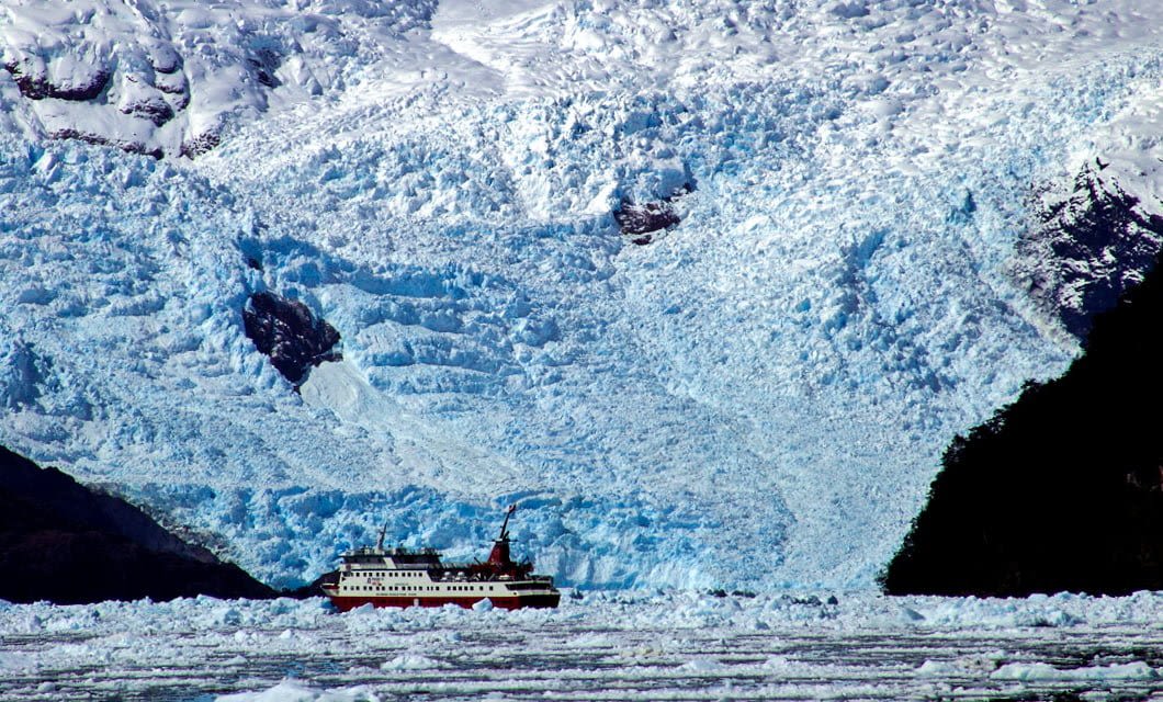 A red and white tour boat from Skorpios Cruises sails near a massive blue glacier in the Chilean Fjords. Snow-covered mountains rise above, while floating ice pieces cover the cold Patagonian waters in the foreground.