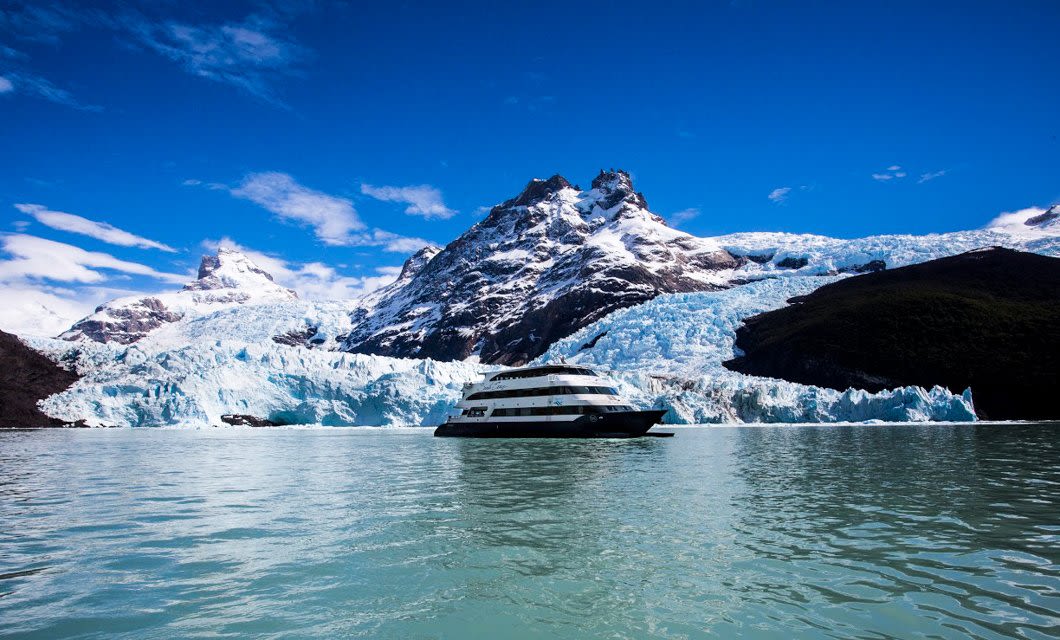 A large Patagonia Deluxe boat sails on turquoise water near a glacier, with snow-covered mountains rising in the background under a bright blue sky.
