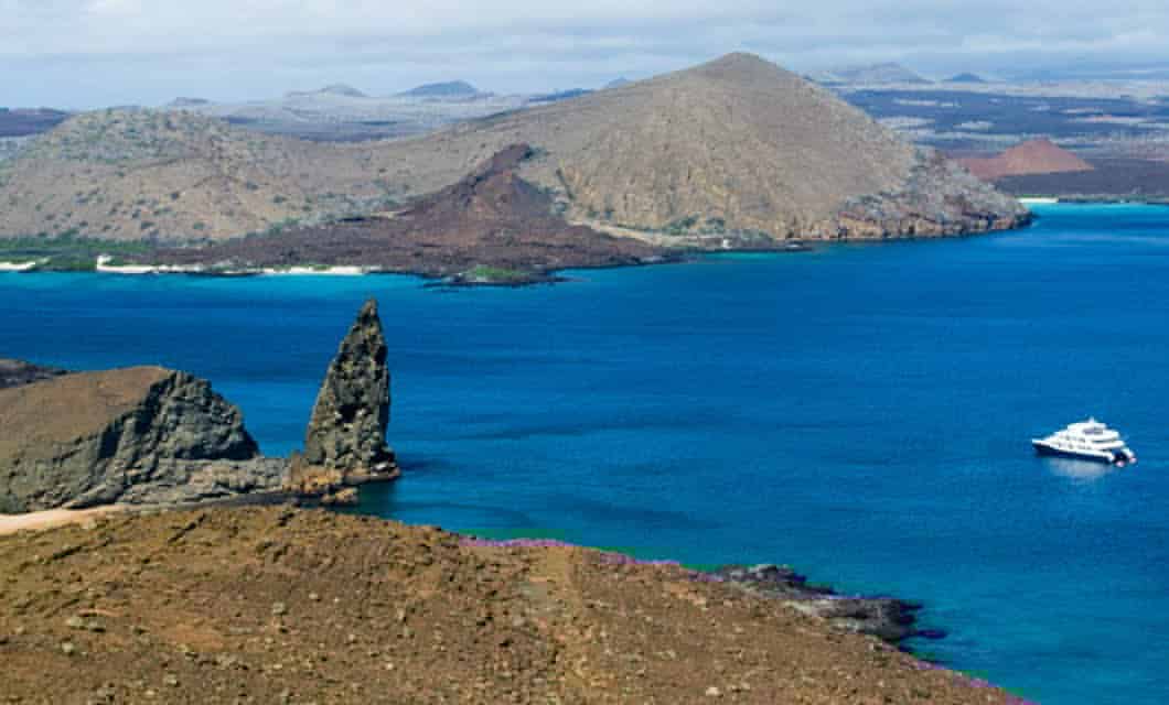 A scenic view of the Galapagos Islands featuring a rugged coastline, a tall rock formation jutting from turquoise waters, a white yacht from the Cormorant cruise, and arid hills in the background under a partly cloudy sky.