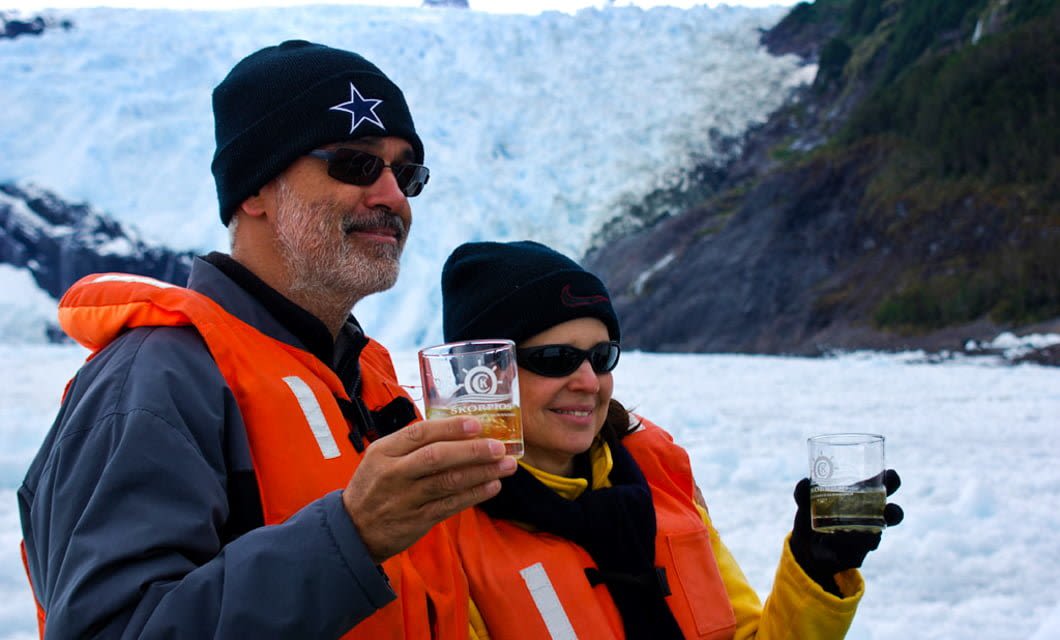Two people wearing orange life vests, black beanies, and sunglasses hold drinks in clear glasses while standing in front of a glacier in the breathtaking Chilean Fjords.