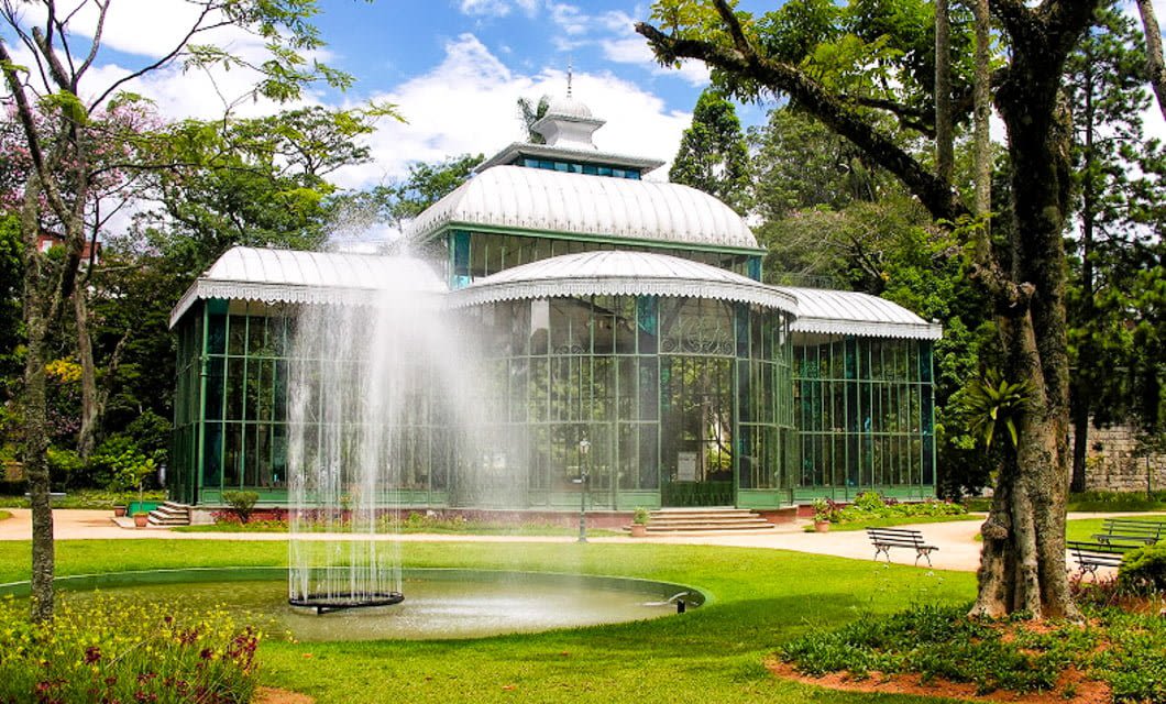 A large glass greenhouse with a metal roof sits behind a circular fountain spraying water, surrounded by green grass, trees, benches, and colorful flowers—a serene stop along the Brazil Gold Route under a partly cloudy sky.