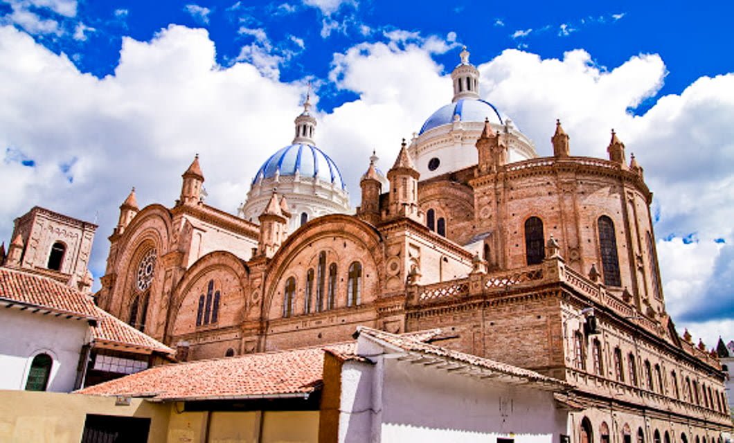 A grand cathedral with large blue and white domes and ornate brickwork stands under a bright Ecuador Tour sky with scattered clouds, seen from a low angle with part of a rooftop in the foreground.