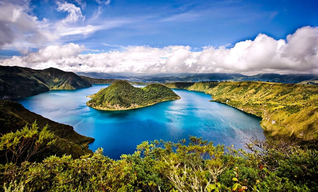 A large blue lake surrounded by lush green hills and mountains under a partly cloudy sky, reminiscent of the breathtaking landscapes seen on a Galapagos Cruise, with a small island and vibrant vegetation in the foreground.