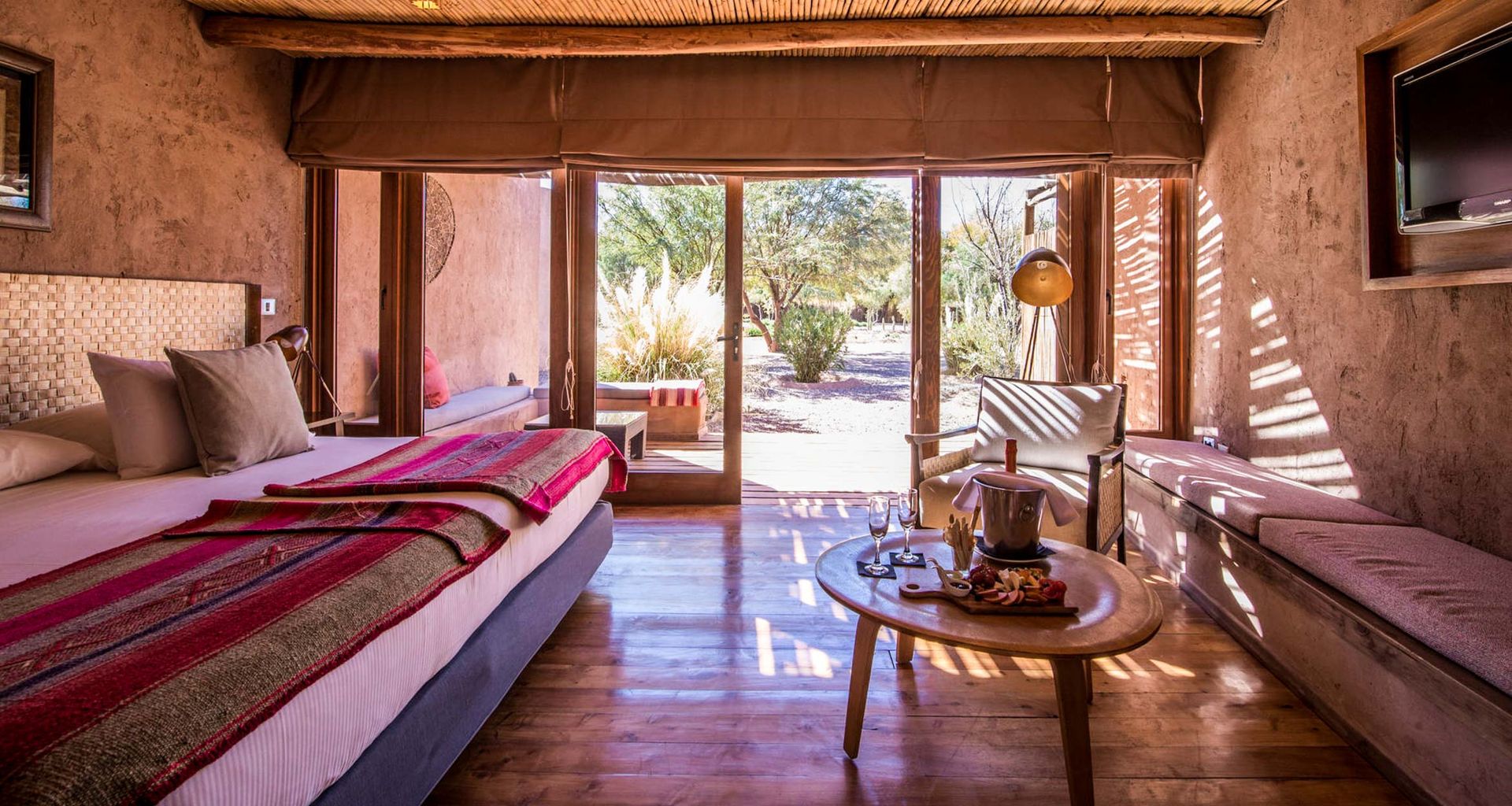 A cozy, sunlit Cumbres Atacama hotel room with a large bed, colorful blanket, wooden floors, and rustic decor. A round table with a drink tray sits by glass doors opening to a patio with desert plants—perfect for relaxation after the spa.