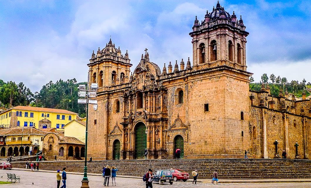 A large historic stone cathedral with two towers stands on a wide plaza, where people from a Bolivia train tour stroll among parked cars. The sky is cloudy, with colorful buildings visible to the left and behind the cathedral.