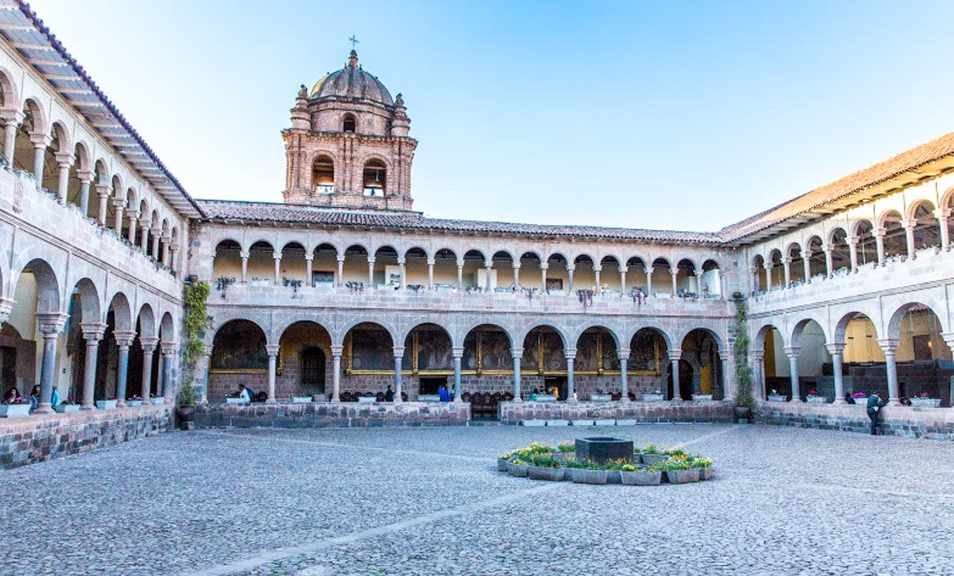 A spacious stone courtyard surrounded by arched two-story colonial cloisters, reminiscent of Machu Picchu’s grandeur, with a prominent dome rising above the building under a clear blue sky.