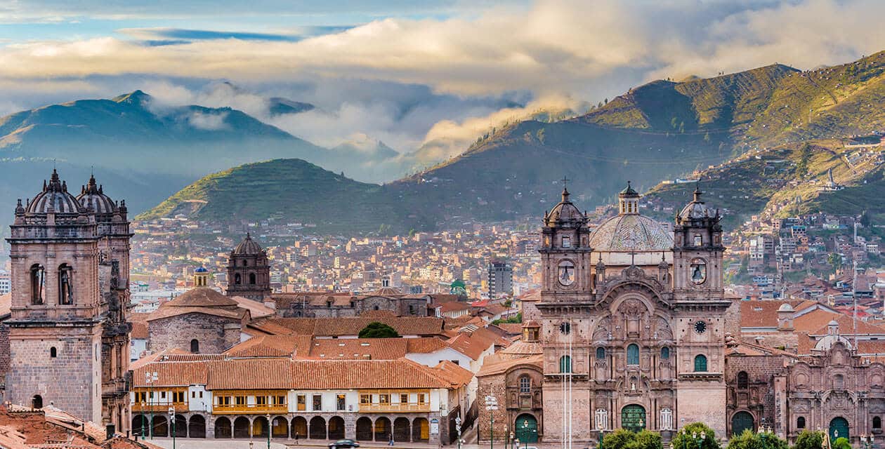Panoramic view of Cusco, one of South America's Cultural Capitals, featuring historic colonial buildings with red-tiled roofs, cathedral towers, and green mountains in the background under a cloudy sky.