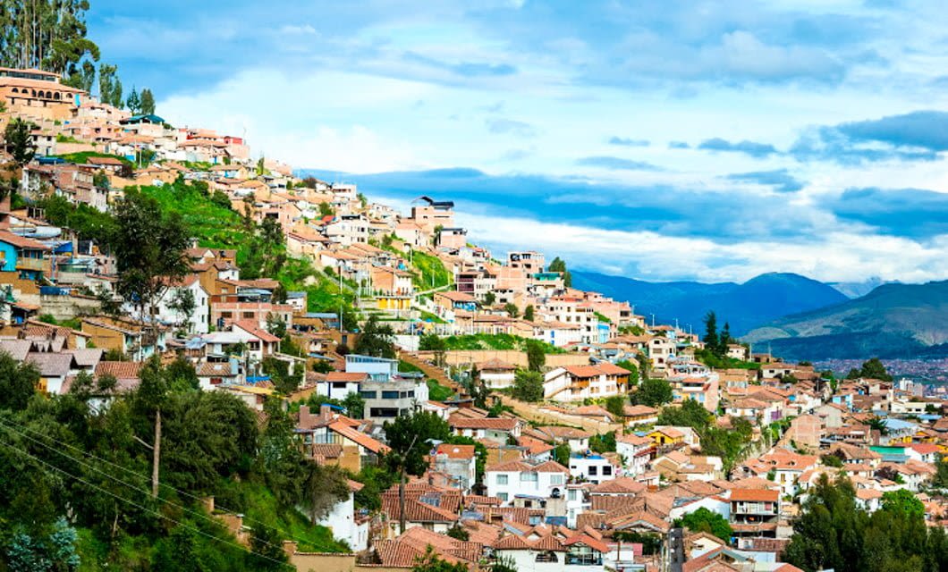 Hillside cityscape with colorful houses and terracotta rooftops nestled among green trees, set against distant mountains and a partly cloudy sky—an inviting stop before your Machu Picchu or Amazon Jungle Tour adventure.