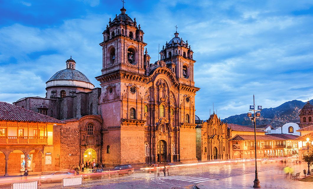 A large, ornate stone church with twin bell towers stands in a lively town square at dusk, surrounded by colonial-style buildings—an inviting scene often found on a Peru Trek, with distant mountains under a blue, cloudy sky.