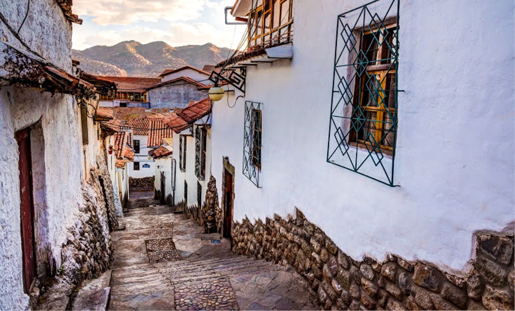 A narrow, cobblestone street lined with white colonial-style buildings and barred windows winds downhill, offering a quintessential South America view with red-tiled roofs and mountains in the background under a partly cloudy sky.