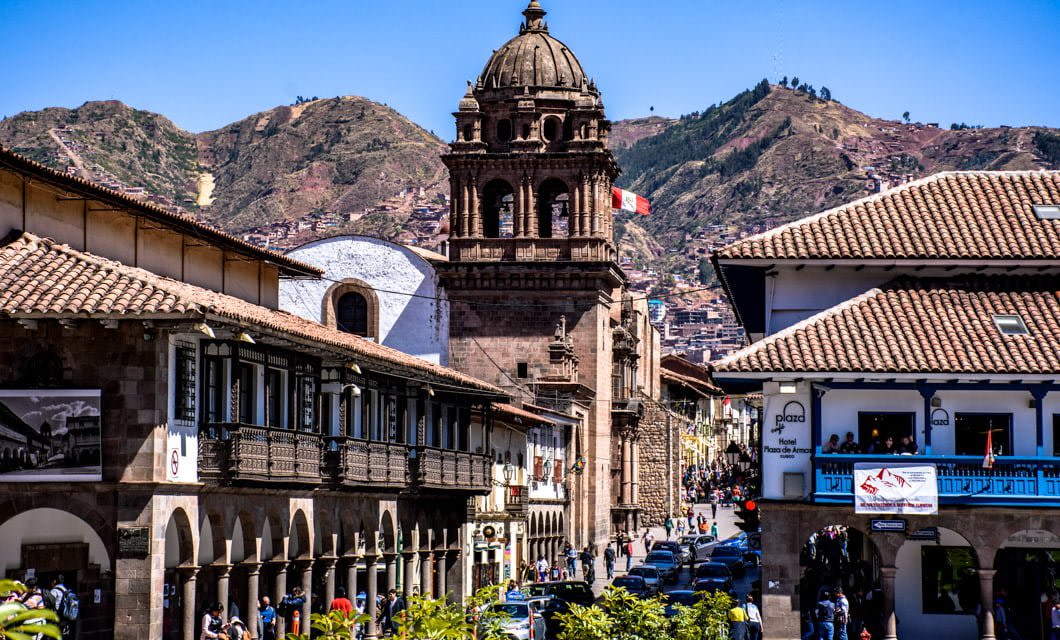 A lively street scene in a historic South American city with colonial buildings, a stone church tower, and mountains beckoning for trekking in the background. People and cars fill the streets as a Peruvian flag waves atop the tower.