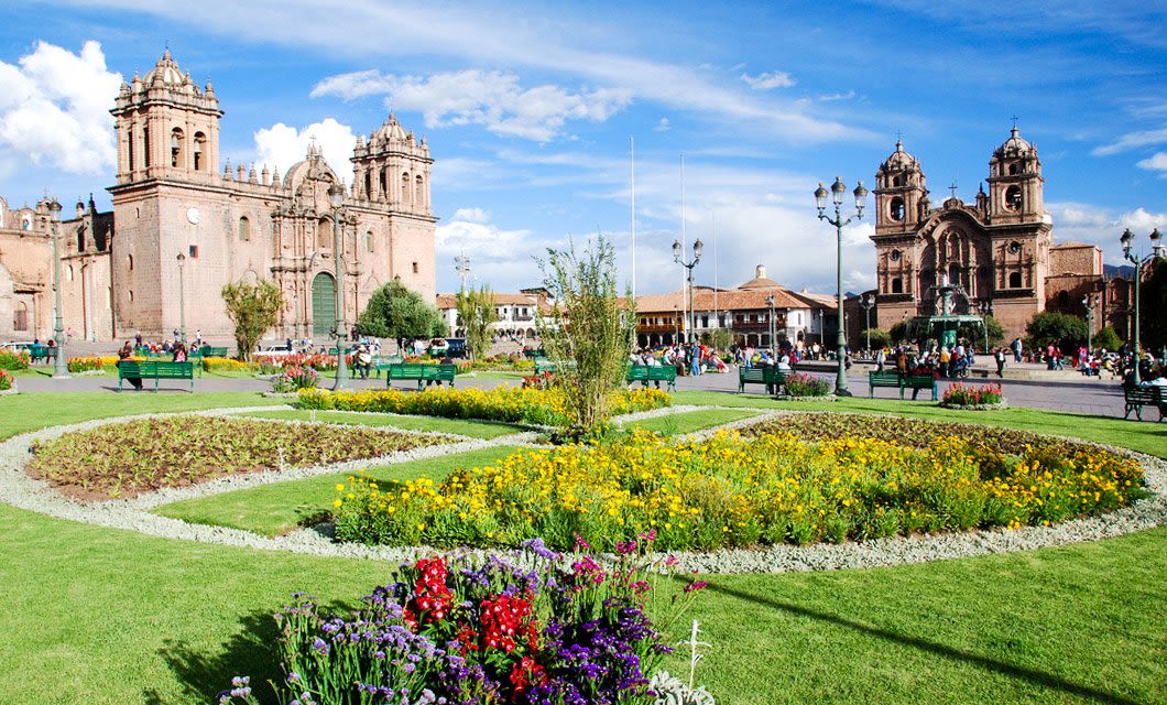 A vibrant city square in La Paz with colorful flower beds, green lawns, benches, and historic stone churches under a blue sky with scattered clouds; people walk and gather in the background, echoing the spirit of the Travels of the Incas.