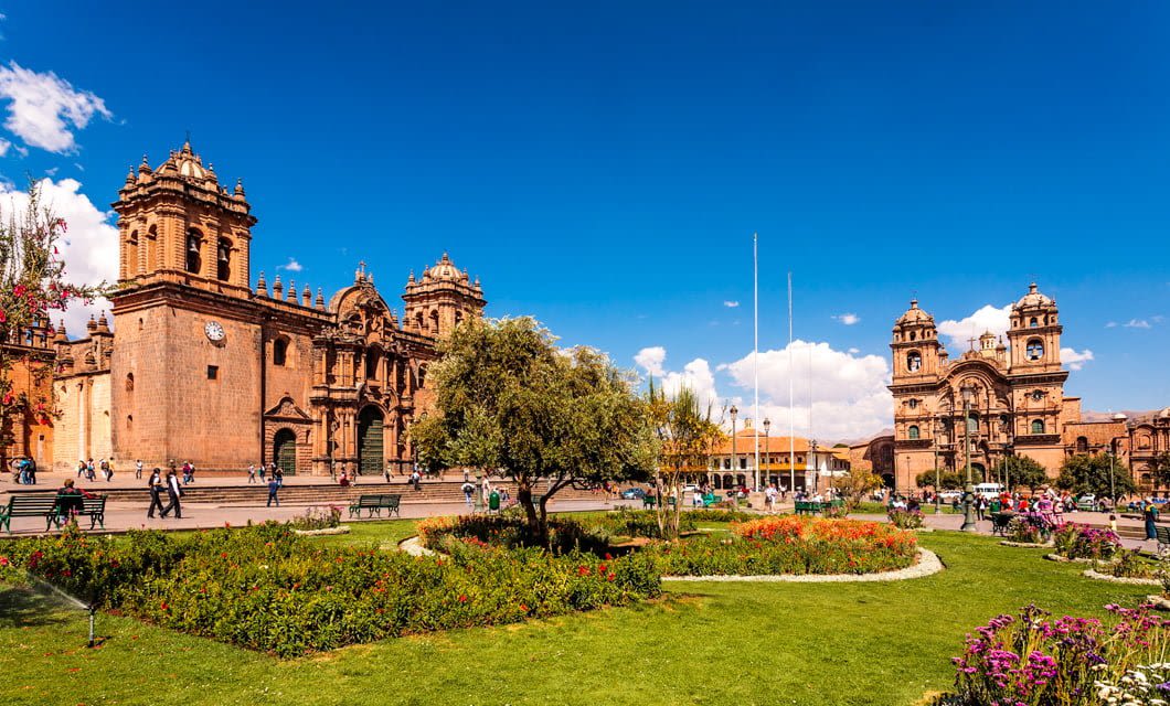 A vibrant plaza in South America with green gardens, benches, and people walking—some resting after trekking nearby trails—surrounded by historic stone buildings with intricate facades under a bright blue sky.