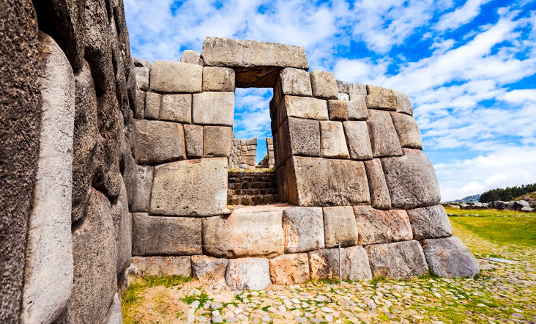 Ancient stone wall with a rectangular doorway at Sacsayhuamán in Peru, part of an unforgettable South America Tour. Large, irregular stones are fitted tightly together under a blue sky, with grass and stone paths surrounding the structure.