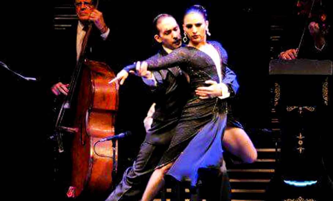 A couple performs an intense tango dance on stage, evoking the spirit of Argentina, with a double bass player and another musician in the background under dramatic lighting.