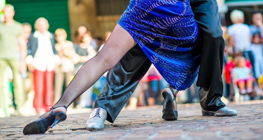 Two dancers perform a passionate tango on a cobblestone street in Argentina; one wears a blue dress and black fishnet stockings, the other black pants. A blurred crowd looks on, soaking up the lively spirit of this food and wine tour.