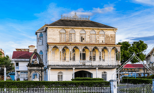A large, historic wooden building with arched windows and intricate decorative trim, surrounded by a white fence and greenery, under a blue sky.