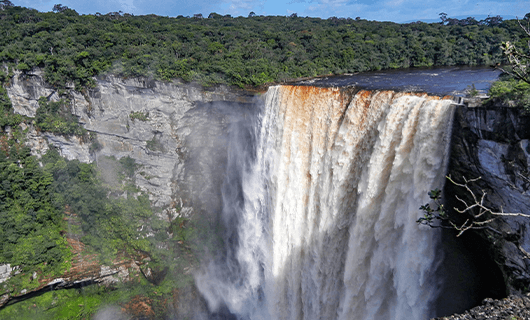 A wide, powerful waterfall cascades over a cliff surrounded by lush green forest under a blue sky, creating mist at the base.